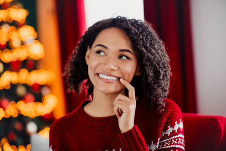 Cheerful woman in red Christmas sweater smiles by a sparkling Christmas tree at home dreaming of merry holiday magicの写真素材