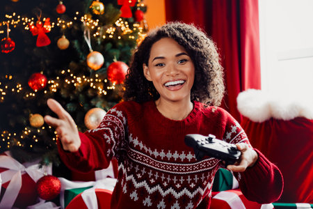 Funny joyful woman in red Christmas sweater reaches for a game controller by a lit Christmas tree at homeの写真素材