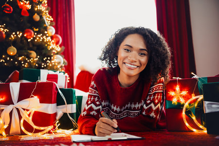 Smiling woman in warm christmas sweater writes on clipboard amid gifts and twinkling tree lights at home for festive joy and holiday planningの写真素材