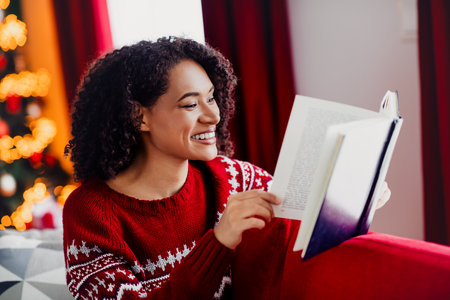 A joyful woman sits on a cozy sofa reading a book during Christmas season in a warm home with a lit tree and festive decorationsの写真素材