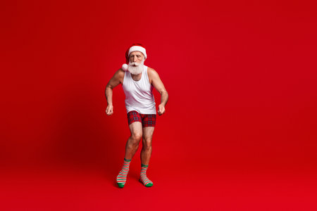 Santa fitness enthusiast dances in a red studio wearing a Santa hat white tank top plaid shorts and striped socks for Christmas joy and holiday energyの写真素材