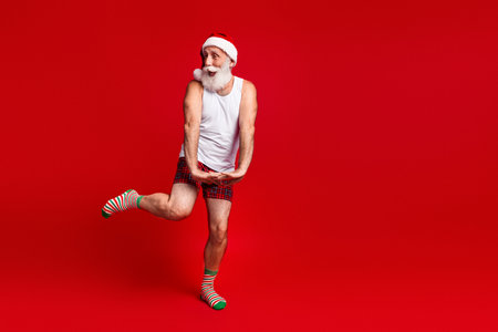 A mature santa look man wearing pajama shorts and santa hat dances playfully against a bold red backdrop for christmas season celebrationの写真素材