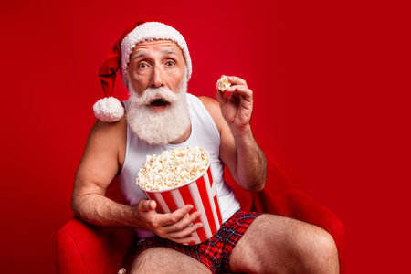 Festive Santa wearing hat and white tank top sits in a bold red room enjoying a large bucket of popcorn during Christmas eve at homeの写真素材