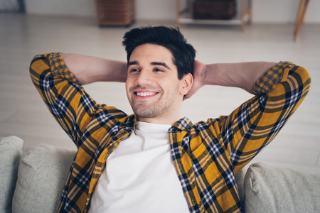 Photo of handsome cheerful guy dressed plaid shirt enjoying weekend relaxing sofa arms behind head indoors room home houseの写真素材