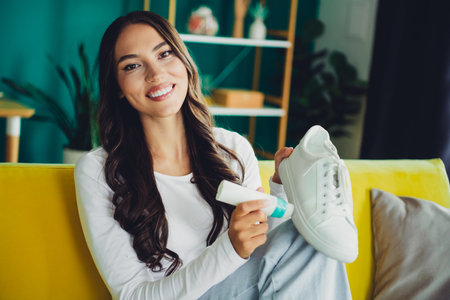 Cheerful latin woman at home polishing white sneakers on a bright living room sofa with plants showing casual lifestyle and chic interior design.の写真素材