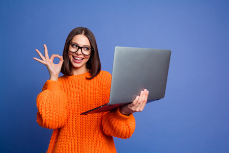 Young woman in an orange sweater gesturing okay while holding a laptop, expressing excitement against a blue backgroundの写真素材