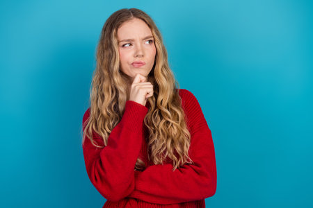 Lovely young woman in red sweater thoughtfully posing against vibrant blue background showcasing cheerful expressionsの写真素材
