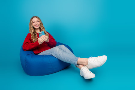 Charming young blonde woman on a blue bean bag chair holding a smartphone, cheerfully relaxing against a vibrant blue backgroundの写真素材