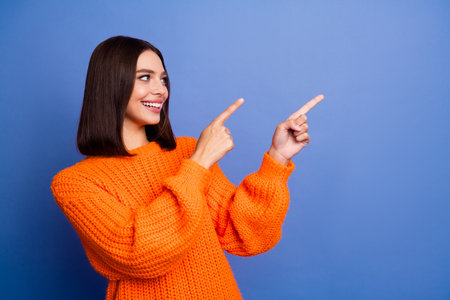 Cheerful young woman in an orange sweater gesturing upwards with both hands against a vivid blue backgroundの写真素材