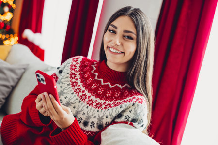 Cheerful woman in red sweater enjoys a cozy Christmas moment at home holding a bright phone with festive decor around herの写真素材