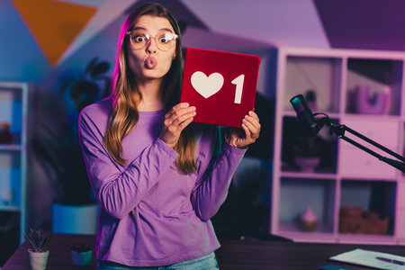 Video chat blogger in casual attire holding a sign with a heart symbol, illuminated by neon lights, engaged in media work at homeの写真素材