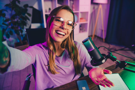 Charming female vlogger recording a podcast in a modern studio illuminated by neon lighting, featuring professional microphone equipmentの写真素材