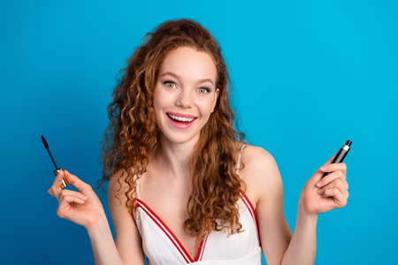 Young cheerful woman with curly red hair holding mascara and lip gloss against blue background for beauty fashion advertising and lifestyleの写真素材