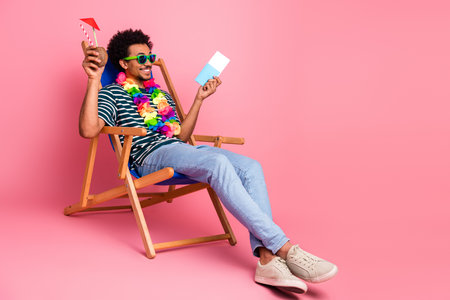 Young mixed race man lounges in a folding chair wearing a lei striped shirt and sunglass reading a card against a pink backgroundの写真素材