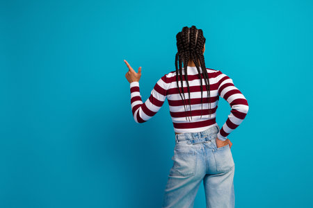 Young stylish woman with braided hair wearing striped top and jeans poses against a bright blue background for fashion lifestyle stock photoの写真素材