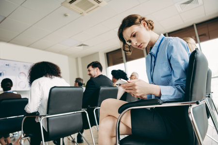 Businesswoman checking phone during conference in modern office space among diverse coworkers focusing on presentationの写真素材