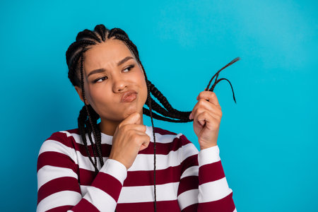 Confident young woman with braids pulls a strand of hair against a bright blue background while...