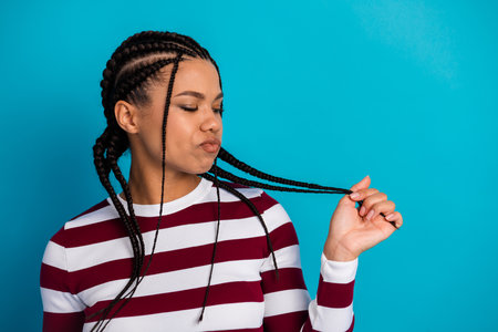 Young woman with braided hair wearing a striped maroon and white top posing against a blue background showing playful hair movement and casual fashionの写真素材