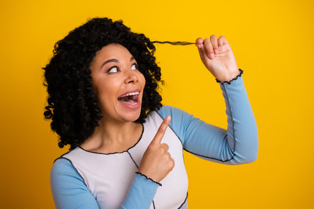 Excited young woman gesturing playfully with her curly hair against a vibrant yellow backdrop expressing joyの写真素材