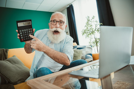 Senior man at home enjoys day working from home with laptop calculator living room featuring interior and comfortable furnitureの写真素材
