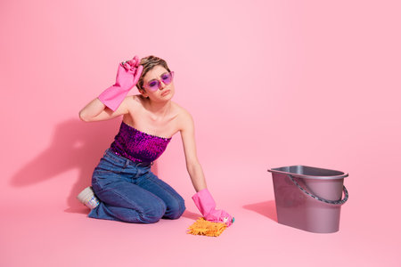 Young glamour woman in pink sequined top and jeans cleaning with mop against pink background with stylish confident vibeの写真素材