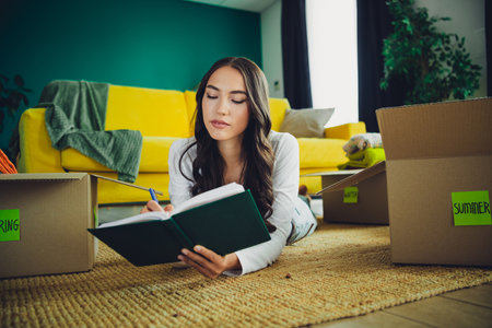 Young woman reads a book on the floor surrounded by moving boxes in a bright living room at home...