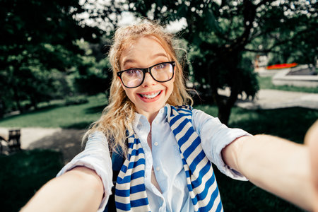 Beautiful woman outdoors taking a selfie in a park, smiling with braces and glasses, showcasing her vibrant lifestyleの写真素材