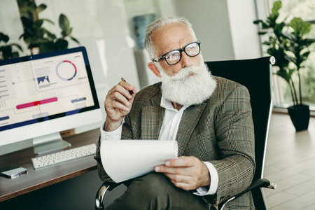 Senior businessman analyzing data with charts on computer monitor in modern office workspaceの写真素材