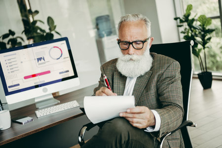 Confident Senior Businessman Reviewing Documents, Analyzing Performance Metrics, and Making Strategic Decisions in Modern Officeの写真素材