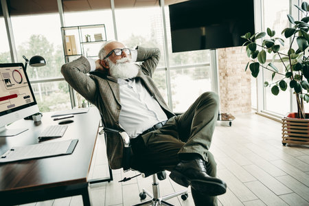 Senior businessman relaxing in a modern office workspace with computers and plantsの写真素材