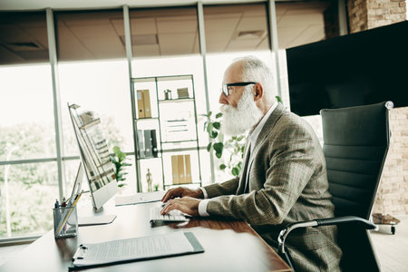 Senior businessman working on a computer in a modern office, analyzing data and planning strategies...