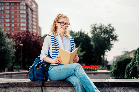 Charming girl reading a book in an urban park with a lively cityscape background during a sunny dayの写真素材