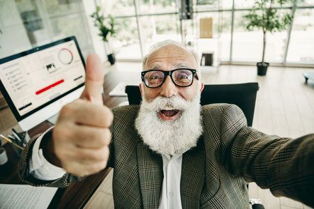 Elderly businessman with thumbs up gesture showcasing confidence and success in an office workspace with a computerの写真素材