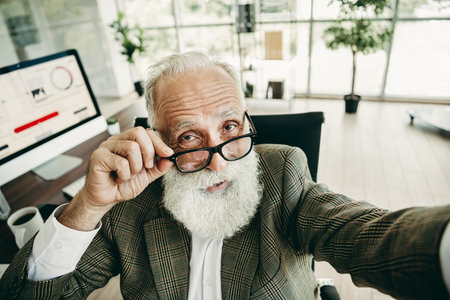 Elegant senior businessman with white beard adjusting glasses in modern office workspace showcasing confidenceの写真素材