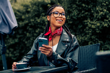 Smiling young woman with phone at outdoor coffee shop wearing a leather jacket and scarf enjoying a sunny dayの写真素材