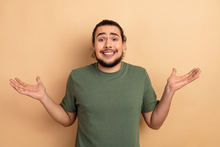 Young man with a shrug and smile wearing a green tshirt stands against a beige background in a casual lifestyle advertising momentの写真素材