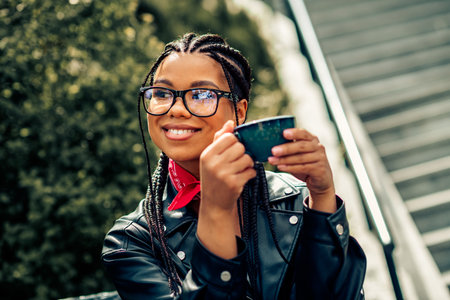 Young woman enjoying leisure outdoors with a cup of tea wearing braided hair, glasses, leather jacket, and scarfの写真素材