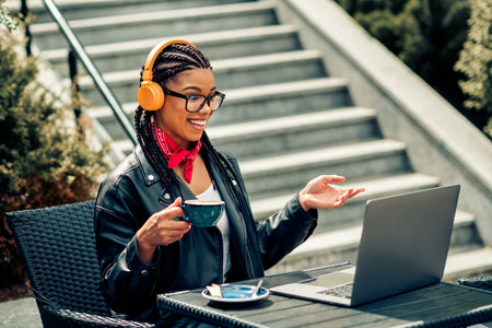 Lovely woman with braided hair enjoying a coffee outdoors while using a laptop and wearing headphones, urban settingの写真素材