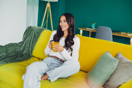 Woman relaxes at home on bright yellow couch enjoying a coffee in a cozy living room sceneの写真素材