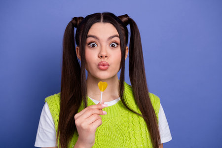 Young female with bright green sweater puckers lips while holding a heart shaped lollipop against a violet backgroundの写真素材