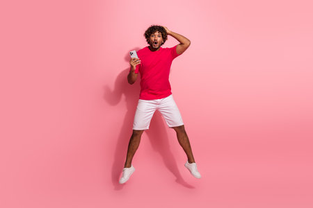 Excited young man with curly hair wearing casual clothes holding smartphone while expressing surprise on a pink backgroundの写真素材