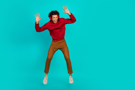 Young man jumps in mid air wearing red sweater brown pants against teal background conveying joy...