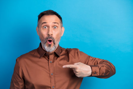 Surprised businessman points to the side during a studio portrait wearing a brown shirt against a vivid blue background for corporateの写真素材