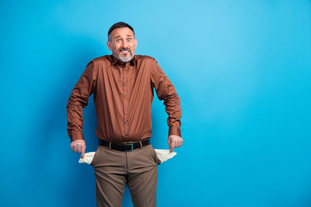 Confident mature businessman with empty pockets smiles in brown shirt against blue background suitable for corporate stock photographyの写真素材
