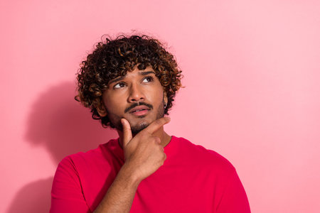 Young man with curly hair thinking thoughtfully while wearing a red t-shirt against a pink backgroundの写真素材