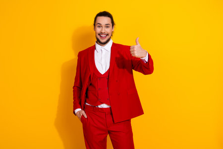 Cheerful man in bold red suit gives thumbs up against bright yellow background for fashion promotionの写真素材