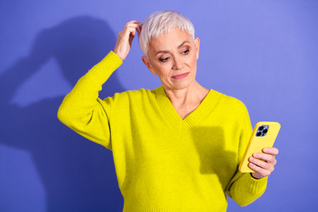 Senior woman in bright yellow sweater using smartphone in purple studio looking thoughtful and stylish fashion lifestyle portraitの写真素材