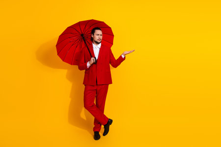 Bold stylish man in red suit carries red umbrella against vibrant yellow background creating a fashion forward lifestyle image with humorous glamorous vibe and modern commercial appealの写真素材