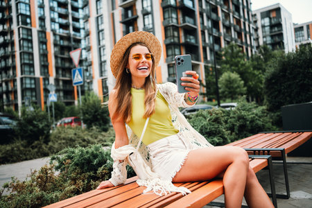 Smiling woman takes a sunny city park selfie wearing a sunhat and bright outfit while enjoying travel vibes and modern styleの写真素材