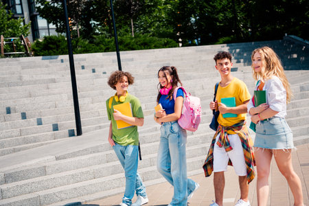 Smiling group of diverse students walking together outdoors during a sunny day holding book and backpacksの写真素材